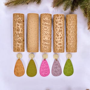 Può includere: Una collezione di mattarelli beige e orecchini a goccia. I mattarelli presentano vari disegni in rilievo, tra cui case, linee e motivi natalizi. Gli orecchini sono nei toni dell'arancione, verde, rosa e viola, con accenti dorati.