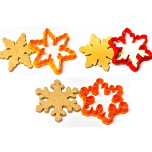 May include: Three snowflake-shaped cookie cutters in orange and red, with baked cookies. The cookies are light brown, and the cutters are plastic. The image is on a white background, showcasing the baking tools.
