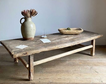 May include: A weathered, rectangular wooden coffee table with a distressed finish. It features a vase with dried flowers, a wooden bowl, and a glass. The table is supported by angled legs and a horizontal beam.