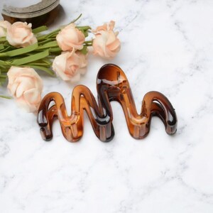 May include: A brown, wavy hair claw clip. The clip is made of a translucent material and has a glossy finish. The clip is designed to hold hair in place and is suitable for various hairstyles. The background includes peach-colored roses and green stems.