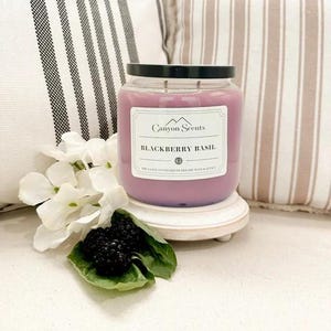 May include: A purple candle with a black lid and a white label that reads "Canyon Scents Blackberry Basil". The candle is sitting on a white wooden pedestal.
