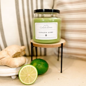 May include: A green glass candle jar with a black lid. The candle is labeled "Canyon Scents" and "Ginger Lime". The candle is on a wooden stand with a lime and ginger root in the foreground.