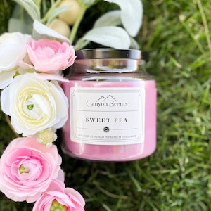 May include: A pink glass candle jar with a black lid. The label reads "Canyon Scents" and "Sweet Pea" with the tagline "The Gold Standard in Premium Fragrance".