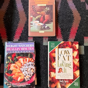 Może przedstawiać: Trzy książki kucharskie z kolorowymi okładkami. "Caribbean Cookery" przedstawia owoce tropikalne. "Weight Watchers Meals in Minutes" pokazuje posiłek z kurczakiem i ryżem. "Low Fat & Loving It" przedstawia makaron.