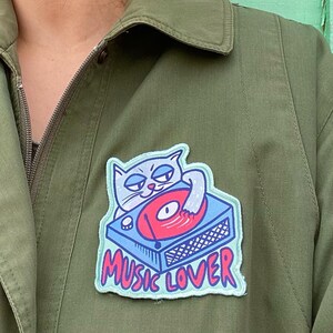 Puede incluir: Una chaqueta verde con un parche bordado en azul y blanco de un gato tocando un disco en un tocadiscos. El parche dice "Music Lover".