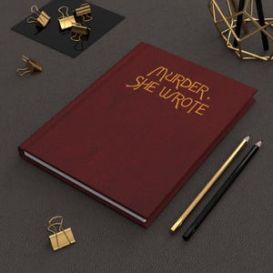 Könnte beinhalten: Ein weinrotes Notizbuch mit dem goldenen Text "MURDER, SHE WROTE". Das Notizbuch ist von goldenen Büroklammern, einem schwarzen und goldenen Bleistift und einem goldenen Bleistift umgeben. Das Notizbuch liegt auf einer dunkelgrauen Oberfläche.