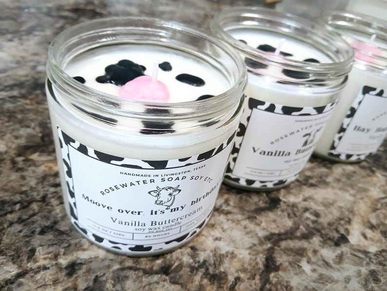 Może przedstawiać: Trzy szklane słoiki z białymi świecami ozdobionymi czarno-białym wzorem krowy. Świece są oznaczone jako "Vanilla Buttercream", "Vanilla Buttercream" i "Hay Day". Świeca na pierwszym planie ma na g&oacute;rze r&oacute;żowo-czarny wz&oacute;r krowy.
