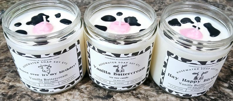 Może przedstawiać: Trzy świece sojowe w szklanych słoikach z czarno-białym wzorem krowy. Świece są oznaczone "Vanilla Buttercream" i mają r&oacute;żową świeczkę urodzinową w środku. Tekst na etykietach brzmi "Move over it's my birthday!" i "Hay Happy Birthday!"