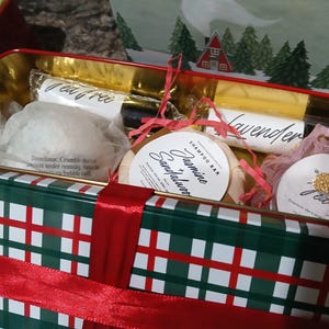 May include: A gift box with a green and red plaid pattern, tied with a red ribbon. Inside are various bath products, including a shampoo bar, bath bombs, and soap bars labeled "Tea Free" and "Lavender". The box has a winter scene on the lid.