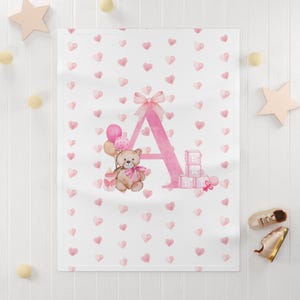 May include: A white baby blanket featuring a repeating pattern of small pink hearts. The center displays a large pink letter A decorated with a bow, a brown teddy bear with a pink ribbon, pink balloons, a baby bottle, and blocks spelling BABY.