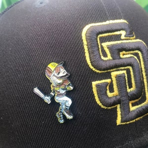 May include: A brown baseball cap with a yellow embroidered 'S' on the front. A baseball player pin is attached to the cap. The pin is of a cartoon baseball player wearing a brown and yellow uniform, holding a bat and looking up.