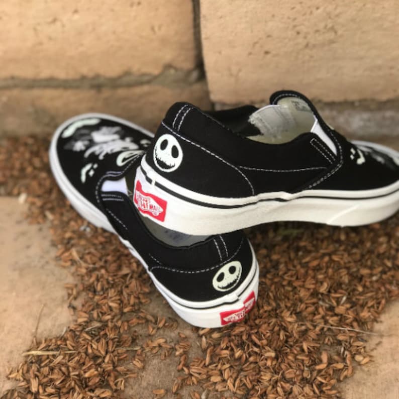 May include: Black slip-on Vans shoes with a white sole and a Jack Skellington graphic on the back. The shoes are on a brown surface.