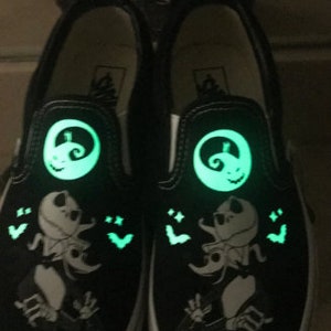 May include: A pair of black slip-on Vans shoes with a glow-in-the-dark design featuring Jack Skellington from The Nightmare Before Christmas. The shoes have a white sole and a green glow.