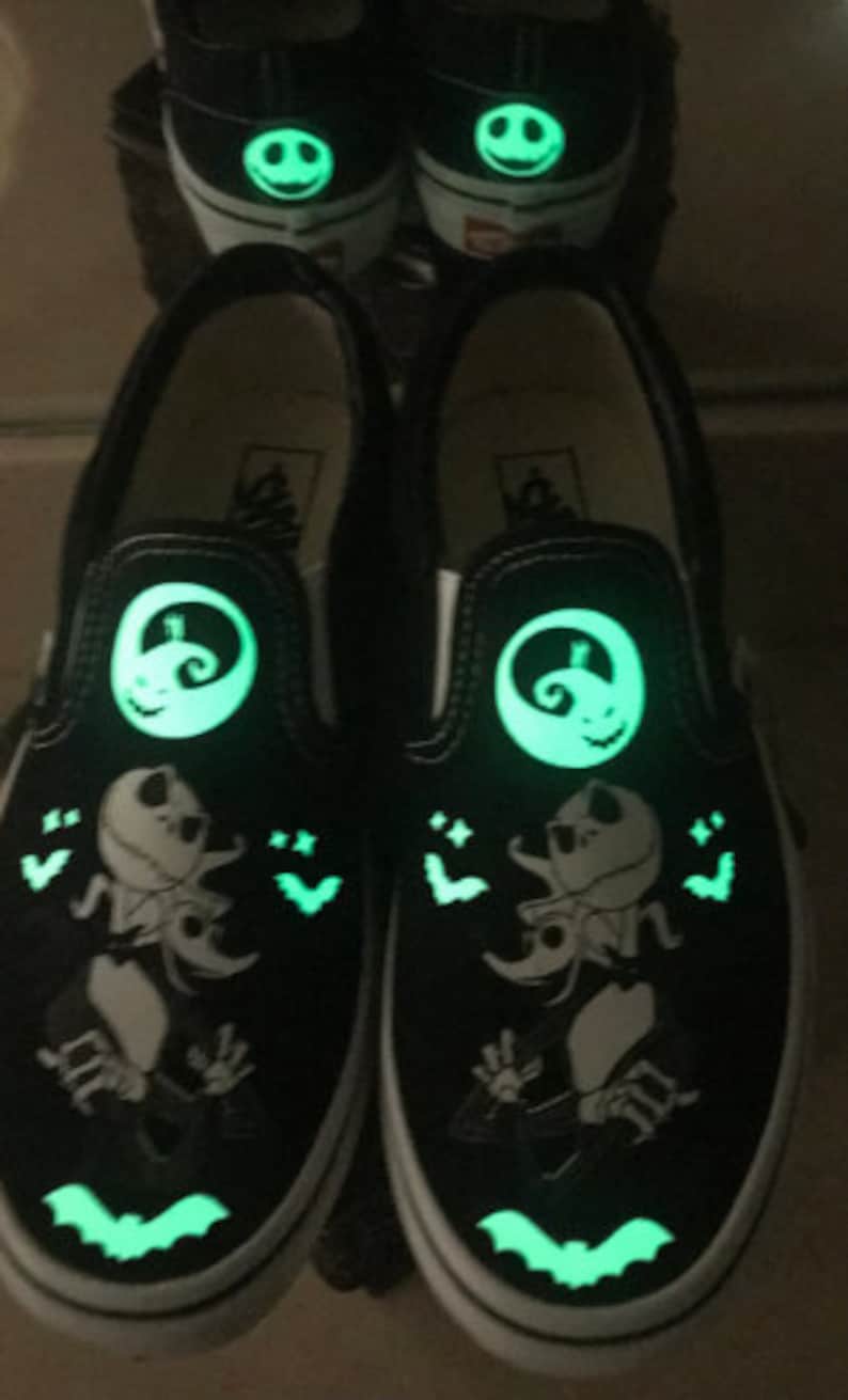 May include: A pair of black slip-on Vans shoes with a glow-in-the-dark design featuring Jack Skellington from The Nightmare Before Christmas. The shoes have a white sole and a green glow.