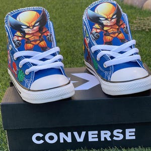 May include: A pair of blue Converse high-top sneakers with a cartoon image of Wolverine on each shoe. The shoes are on a black box with the word "CONVERSE" printed in white.