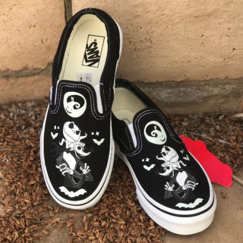 May include: Black slip-on Vans shoes with a glow-in-the-dark design featuring Jack Skellington from The Nightmare Before Christmas. The shoes have a white sole and a black canvas upper.