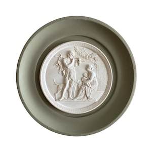 May include: A round, off-white relief sculpture depicting a classical scene of figures and a dog, set within a sage green circular frame. The relief shows a man with a flute, a woman, and a child. The frame has a matte finish.