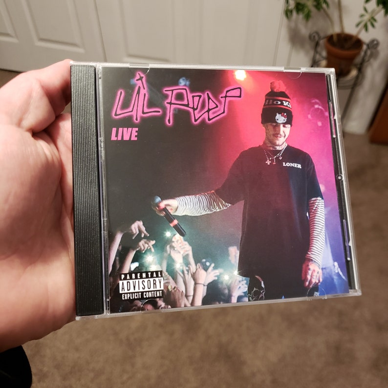 Puede incluir: Una caja de CD con fondo negro y una imagen rosa y roja de Lil Peep. El texto "LIVE" est&aacute; impreso en blanco debajo de la imagen. La imagen muestra a Lil Peep con una camiseta negra con la palabra "LONER" impresa en ella, una camiseta a rayas blancas y negras debajo y una gorra Hello Kitty blanca y negra. La caja de CD tiene una pegatina de advertencia para padres.