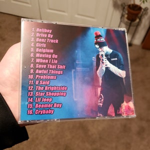 Puede incluir: Una caja de CD con un dise&ntilde;o rosa y azul que presenta a una persona con una camisa a rayas blancas y negras y un gorro rojo y blanco. El texto en la caja enumera los t&iacute;tulos de las canciones: Hellboy, Drive By, Benz Truck, Girls, Belgium, Moving On, When I Lie, Save That Shit, Awful Things, Problems, U Said, The Brightside, Star Shopping, Lil Jeep, Beamer Boy, Crybaby.