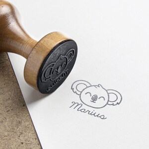May include: A wooden handle rubber stamp with a circular base featuring a koala face design and the name Marius in cursive script. The stamp is shown pressed onto a white surface, leaving a clear black ink impression of the koala and the name.