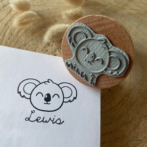 May include: A round wooden rubber stamp featuring a koala face design. The stamp is shown next to a white card printed with the black ink koala graphic and the name Lewis in a cursive script, demonstrating the stamp's clear and precise impression.