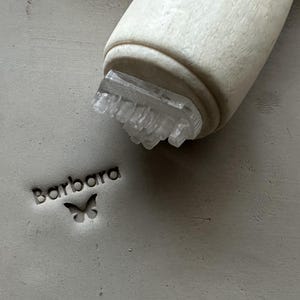 Könnte beinhalten: Ein heller Holzstempel mit einer Basis aus klarem Acryl ist in eine glatte, graue Tonoberfläche gedrückt. Der Stempel hinterlässt einen scharfen, eingeprägten Abdruck des Namens Barbara in einer serifenlosen Schrift sowie ein kleines Schmetterlingssymbol.