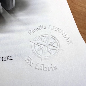 May include: A white embossed design on paper with the words "Famille Lesniak" and "Ex Libris" surrounding a compass rose. The name "N MICHEL" is also visible.