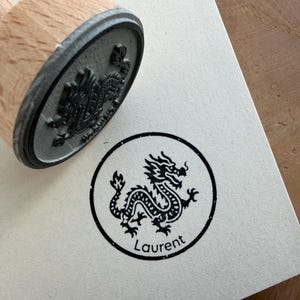 Könnte beinhalten: Ein Holzstempel mit einem schwarzen Drachendesign und dem Namen "Laurent" in einem Kreis gedruckt. Der Stempel ist auf eine weiße Papieroberfläche gedrückt. Der Holzgriff des Stempels ist sichtbar.