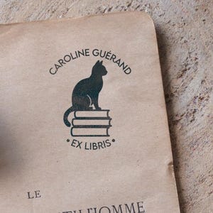 Könnte beinhalten: Ein Stempeldesign mit einer schwarzen Katzen-Silhouette auf drei Büchern. Der Text "CAROLINE GUÉRAND" ist über der Katze, darunter "EX LIBRIS". Der Stempel befindet sich auf strukturiertem, beigefarbenem Papier.