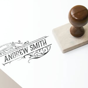 Pode incluir: Um carimbo de borracha preto e branco com o texto "Ex Libris Andrew Smith" em uma fonte decorativa. O carimbo está em uma superfície branca ao lado de uma alça de madeira marrom.