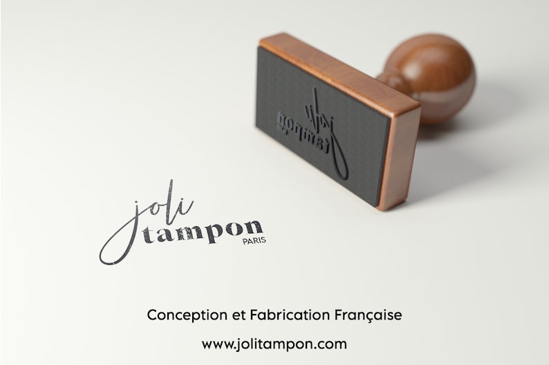 May include: A rectangular wooden rubber stamp with a dark brown handle and a black base. The stamp features the text 'joli tampon PARIS' in a stylized script and serif font. The same text is clearly imprinted in black ink on the surface below.