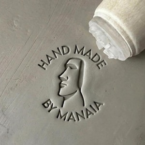 Peut inclure: Un tampon en caoutchouc circulaire avec un manche en bois, pressé sur une surface grise lisse. Le tampon laisse une empreinte représentant le profil d'une tête Moaï stylisée entourée du texte HAND MADE BY MANAIA en police sans empattement.