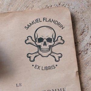 Könnte beinhalten: Ein Holzstempel mit einem Totenkopf- und gekreuzten Knochen-Design, mit dem Text "SAMUEL FLANDRIN" und "EX LIBRIS" in schwarzer Tinte auf braunem Papier gedruckt. Im Hintergrund ist eine Feder zu sehen.