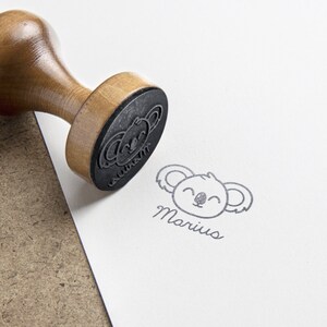 May include: A wooden rubber stamp with a round handle and a black rubber base featuring a koala face design. The stamp is shown next to a clear black ink impression on white paper that displays a koala face and the name Marius in script font.