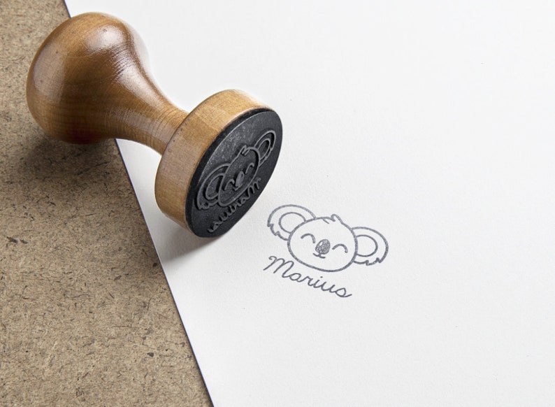 May include: A wooden rubber stamp with a round handle and a black rubber base featuring a koala face design. The stamp is shown next to a clear black ink impression on white paper that displays a koala face and the name Marius in script font.