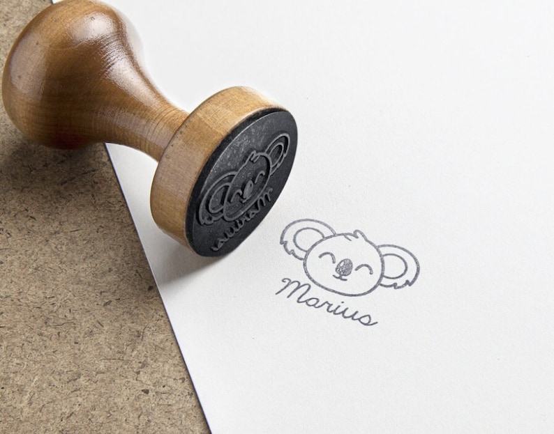 May include: A wooden handle rubber stamp with a circular base featuring a koala face design and the name Marius in cursive script. The stamp is shown pressed onto a white surface, leaving a clear black ink impression of the koala and the name.