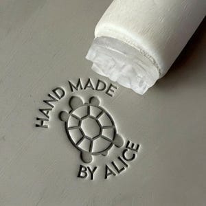 May include: A gray surface with the text "HAND MADE BY ALICE" surrounding a turtle design. A white cylindrical object is partially visible in the upper right corner, suggesting a stamp or tool used to create the impression.