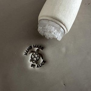 Könnte beinhalten: Ein Holzstempel mit Schildkrötenmuster wird in Ton gepresst und hinterlässt einen Abdruck mit der Aufschrift "HAND MADE BY ALICE". Der Stempel hat eine transparente, strukturierte Basis. Der Ton ist von neutraler grauer Farbe.