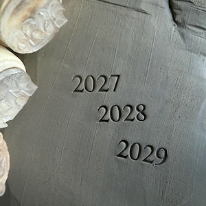 Op de afbeelding: Drie doorzichtige cilindrische stempels met getextureerde uiteinden zijn naast een grijs kleioppervlak geplaatst. De klei bevat de ingedrukte cijfers 2027, 2028 en 2029, elk duidelijk in het materiaal gestempeld in een verticale opstelling.