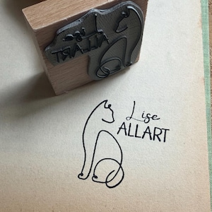 Könnte beinhalten: Ein Holzstempel mit einem Katzendesign und dem Text "Lise ALLART" auf einer beigefarbenen Oberfläche. Der Stempel besteht aus Holz und hat einen Gummistempel eines Katzenmotivs. Das Katzendesign ist ebenfalls auf der Oberfläche abgebildet.