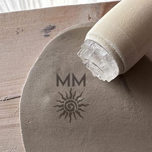 Könnte beinhalten: Ein hellbeige, runder, strukturierter Steinstempel mit den Buchstaben "MM" und einem Sonnendesign. Ein hellbeiger Holzgriff ist an einem transparenten, strukturierten Stempelkopf befestigt. Der Stempel befindet sich auf einer hellbeigen Holzoberfläche.