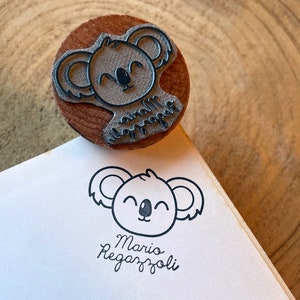 May include: A round wooden rubber stamp featuring a carved koala face and the text Mario Regazzoli. The stamp is shown with a black ink impression of the same koala design and the name Mario Regazzoli printed clearly on a white paper surface.