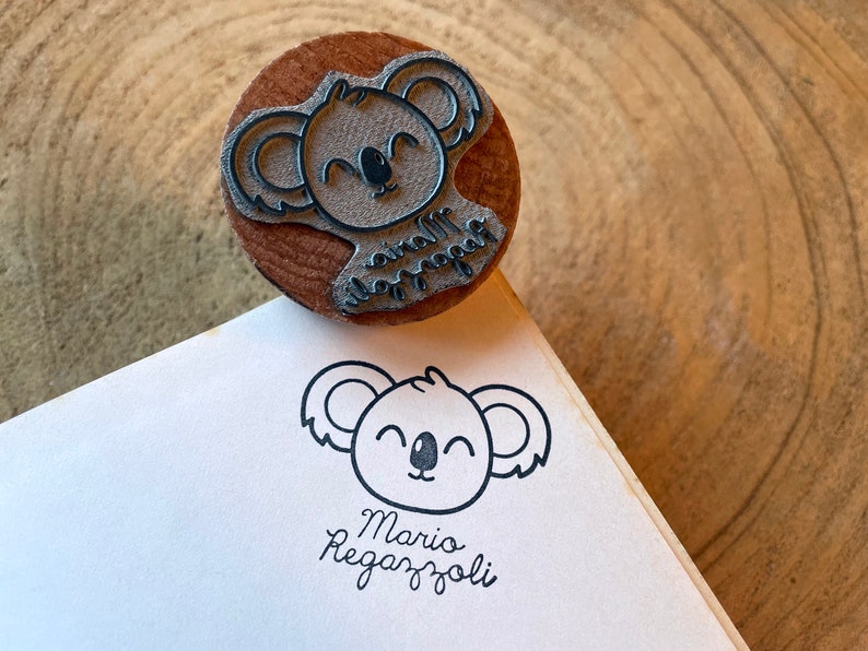 May include: A round wooden rubber stamp featuring a carved koala face and the text Mario Regazzoli. The stamp is shown with a black ink impression of the same koala design and the name Mario Regazzoli printed clearly on a white paper surface.