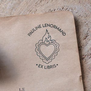 Può includere: Un timbro su carta beige con la scritta "PAULINE LENORMAND" sopra un disegno a forma di cuore con una fiamma e le parole "EX LIBRIS" sotto. Un manico di timbro in legno è visibile a sinistra.