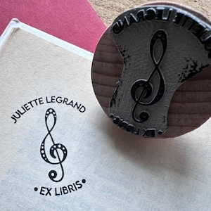 May include: A wooden stamp with a metal imprint of a treble clef, next to a book page with the same design and the text "JULIETTE LEGRAND EX LIBRIS". The stamp is on a wooden surface, and the book is on a red surface.