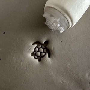 Peut inclure: Un petit tampon en forme de tortue brun foncé pressé sur une surface d'argile grise. Le tampon est détaillé avec un motif de carapace et se trouve à côté d'un outil en plastique blanc et transparent.