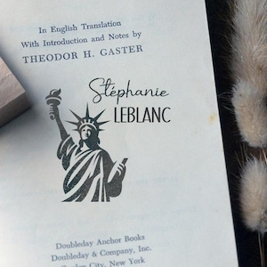 Può includere: Una pagina di libro vintage con un'illustrazione della Statua della Libertà e il nome "Stéphanie LEBLANC". Il testo "In English Translation" e il nome dell'autore sono visibili. Un timbro di legno è nell'angolo in alto a sinistra.