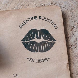 Può includere: Un timbro in legno con un disegno di labbra e il testo "VALENTINE ROUSSEAU" e "EX LIBRIS" stampato su un pezzo di carta marrone chiaro. Una piuma è visibile sullo sfondo.