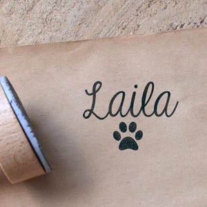 Könnte beinhalten: Ein Holzstempel mit dem Namen "Laila" in Schreibschrift und einem Pfotenabdruck-Symbol. Der Stempel ist auf braunes Papier gedrückt und erzeugt ein personalisiertes Design.