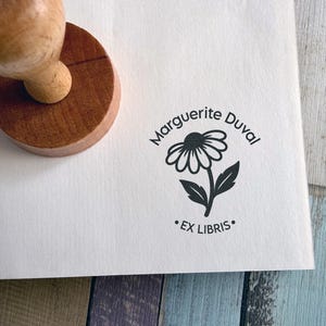 Op de afbeelding: Een houten rubberstempel met een ronde handgreep, die een zwart inktontwerp op wit papier drukt. Het ontwerp toont een madeliefje met de tekst Marguerite Duval in een boog erboven en de woorden EX LIBRIS in het midden eronder.